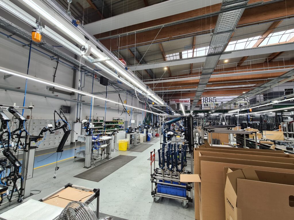 Eine geräumige, moderne Fabrikhalle mit hohen Decken, freiliegenden Balken, Montagestationen, Maschinen, Werkzeugen, Regalen und großen Kartons. Deckenleuchten erhellen den sauberen, organisierten Arbeitsbereich.