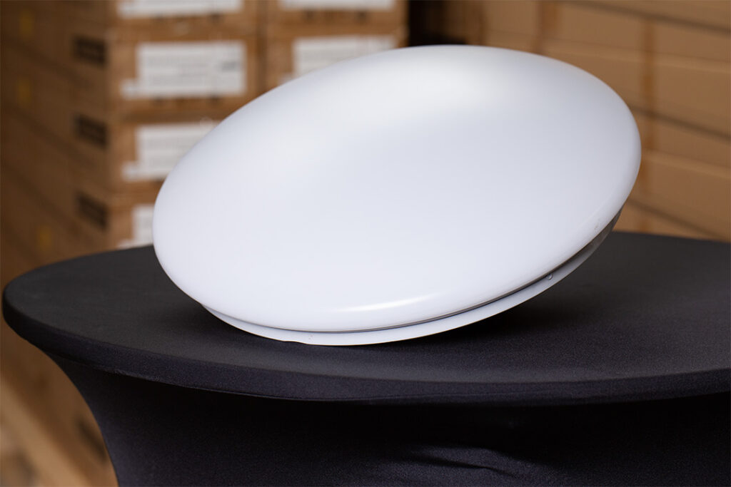 Auf einer schwarzen Tischdecke steht ein weißes, scheibenförmiges elektronisches Gerät, im Hintergrund sind verschwommen gestapelte Pappkartons zu sehen.