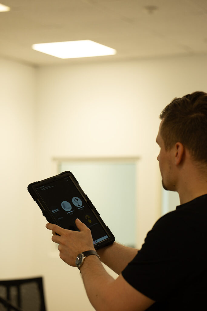 Ein Mann in einem schwarzen Hemd hält und verwendet ein Tablet-Gerät in einem hellen, modernen Büroraum mit einer hellen Wand und einem Fenster im Hintergrund.