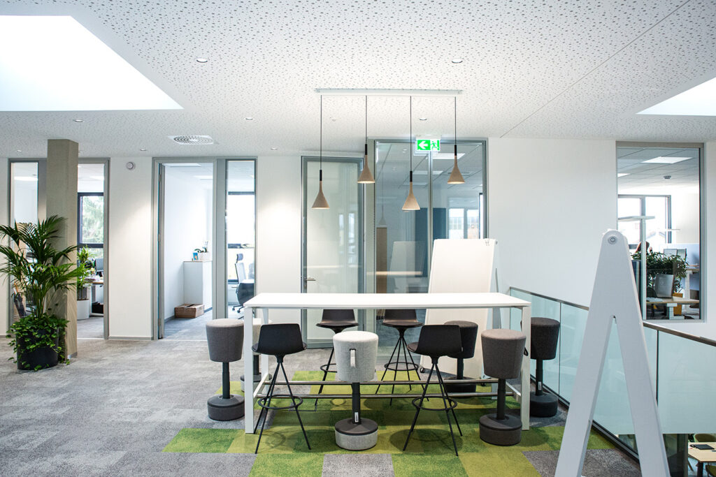 Moderner Büroraum mit einem Stehtisch, umgeben von Hockern, Hängelampen, Pflanzen, Glaswänden und hellem Tageslicht durch Fenster und Oberlichter. Rechts daneben steht ein Whiteboard.