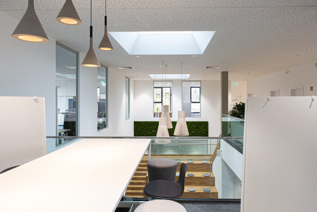Moderner, heller Büroraum mit weißen Schreibtischen, Pendelleuchten, Glasgeländer und einem Oberlicht. Im Hintergrund sind grüne Mooswände und große Fenster zu sehen, die natürliches Licht hereinlassen.
