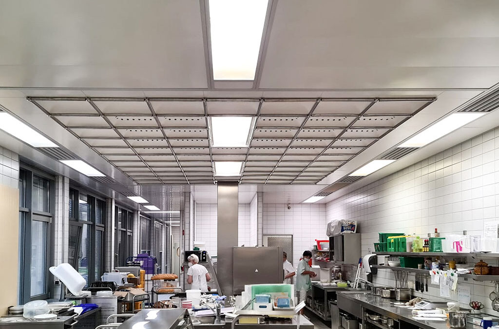 Eine geräumige Profiküche mit Geräten und Arbeitsflächen aus Edelstahl, weiß gefliesten Wänden und Deckenleuchten mit LED-Beleuchtungssystemen. Man sieht zwei Köche bei der Arbeit, einen in der Mitte und einen im Hintergrund. Auf den Arbeitsflächen sind verschiedene Küchenutensilien und Zutaten ausgebreitet.