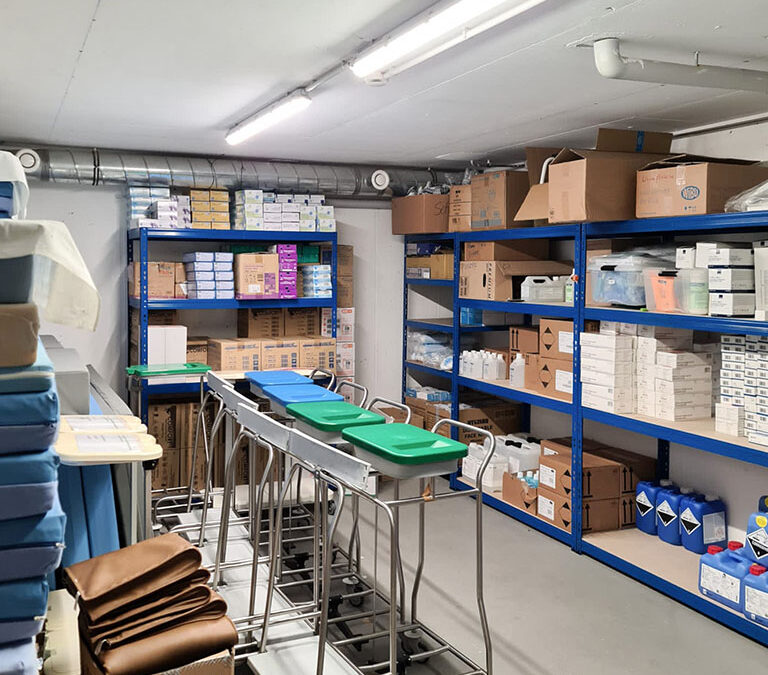 Ein gut organisierter Lagerraum mit Metallregalen, in denen verschiedene medizinische Vorräte, darunter Kisten und Pakete, aufbewahrt werden. Mehrere Metallwagen mit grünen Tabletts stehen in der Mitte des Raums, der mit modernen LED-Beleuchtungssystemen gut beleuchtet ist.
