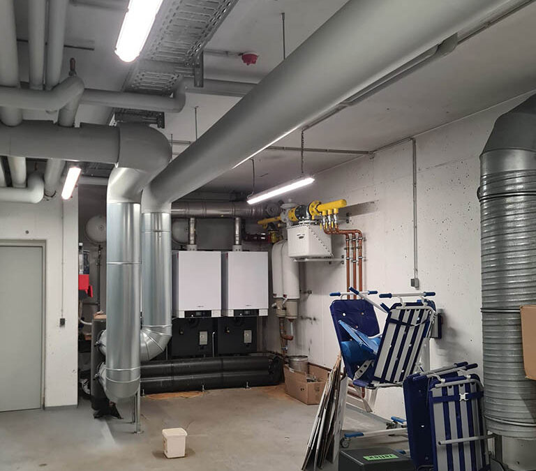 Ein Wartungsraum mit verschiedenen Industrierohren an Decke und Wänden, einem großen Gerät in der Ecke und zwei blauen Klappstühlen, die an der Wand lehnen. Ein Karton und andere kleine Gegenstände liegen verstreut auf dem Boden. LED-Beleuchtung eines erstklassigen Leuchtenherstellers erhellt den Raum.