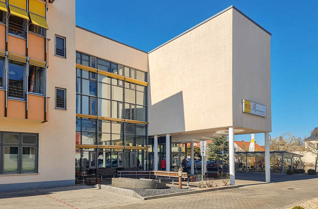 Ein modernes Gebäude mit großen Fenstern und einer beigen Außenfassade. An der Ecke des höheren Gebäudeteils ist ein Schild mit der Aufschrift „BMIB“ angebracht. Der untere Teil, der mit einem hochmodernen LED-Lichtsystem ausgestattet ist, verfügt über einen Eingang mit Glasfassade und Bänken in der Nähe. Im Vordergrund ist ein von Bäumen und Sträuchern gesäumter Weg unter einem klaren blauen Himmel zu sehen.
