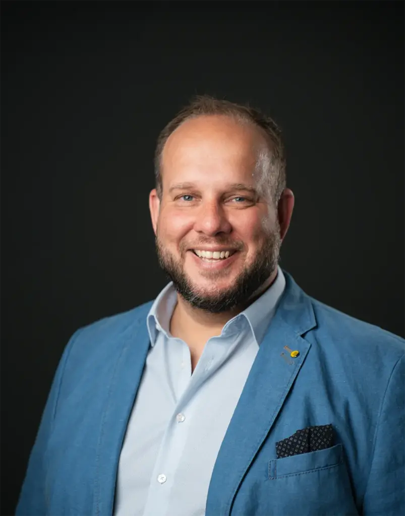 Ein lächelnder Mann mit kurzen Haaren und einem Bart trägt ein hellblaues Hemd und einen blauen Blazer mit einem Einstecktuch und steht vor einem dunklen Hintergrund.
