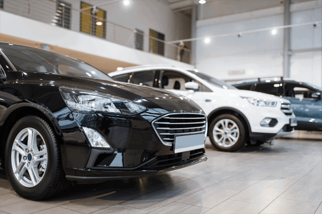 Eine Reihe moderner Autos, darunter eine schwarze Limousine und weiße SUVs, sind in einem hell erleuchteten Autohaus-Ausstellungsraum mit großen Fenstern und gefliesten Böden ausgestellt.
