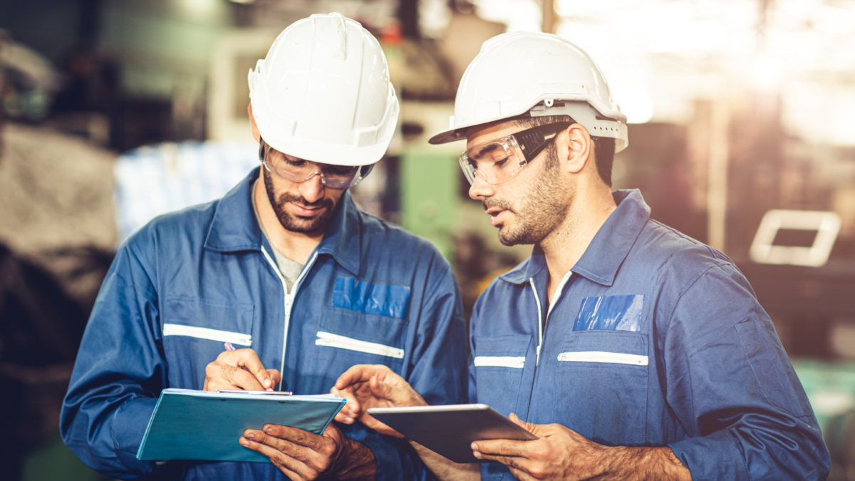 Zwei männliche Arbeiter in blauen Overalls und weißen Schutzhelmen stehen zusammen in einer Fabrik und diskutieren über Sicherheits- und Arbeitsstättenbeleuchtungsnormen. Der eine schreibt auf einem Klemmbrett, der andere hält eine Tafel; beide tragen eine Schutzbrille.