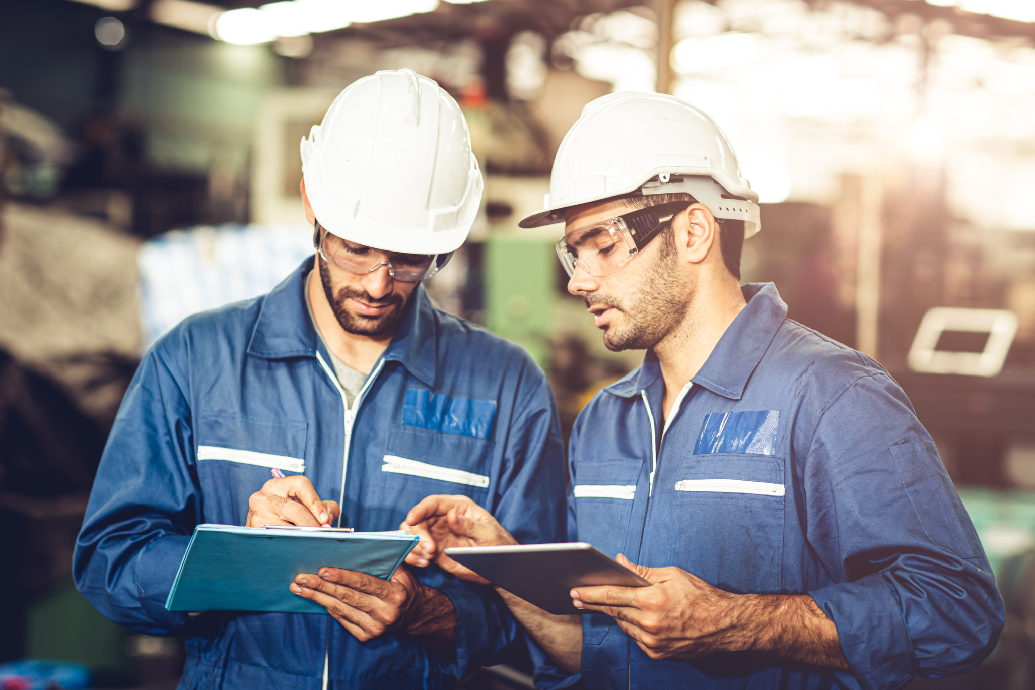 Zwei männliche Arbeiter in blauen Overalls und weißen Schutzhelmen stehen zusammen in einer Fabrik und diskutieren über Sicherheits- und Arbeitsstättenbeleuchtungsnormen. Der eine schreibt auf einem Klemmbrett, der andere hält eine Tafel; beide tragen eine Schutzbrille.