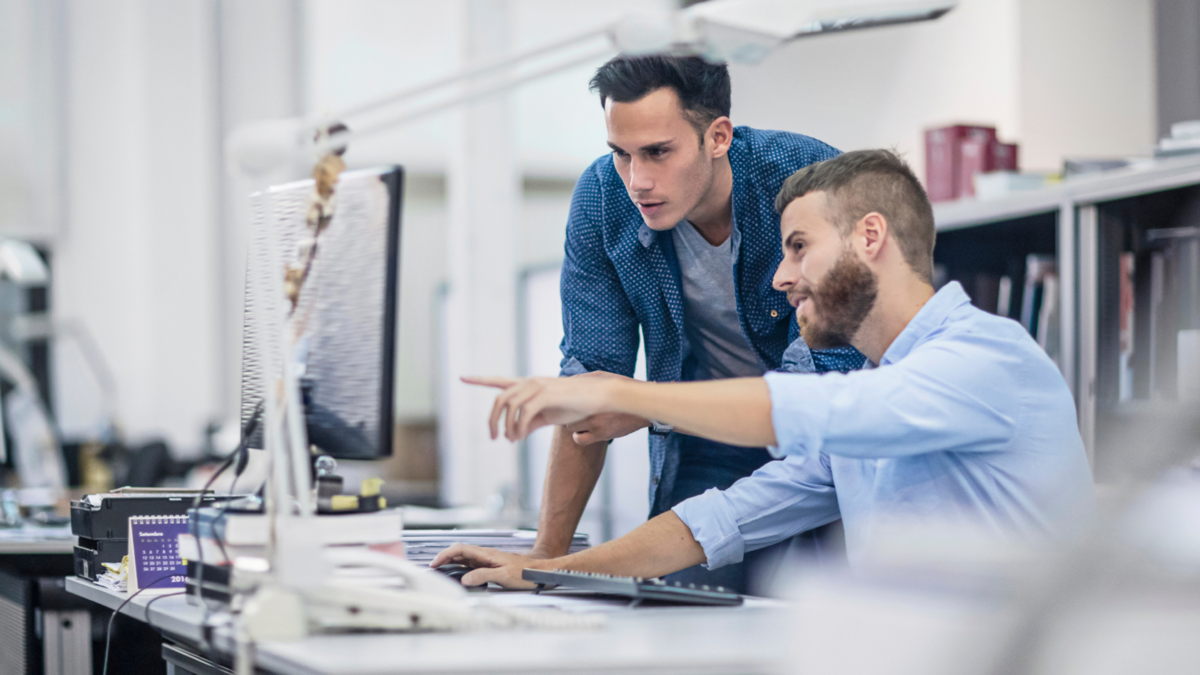 Zwei Männer in einem modernen Büro schauen konzentriert auf einen Computerbildschirm und diskutieren über Lichtplanung. Der eine sitzt und zeigt auf den Monitor, der andere steht daneben. Beide wirken konzentriert und in ihre Arbeit vertieft.