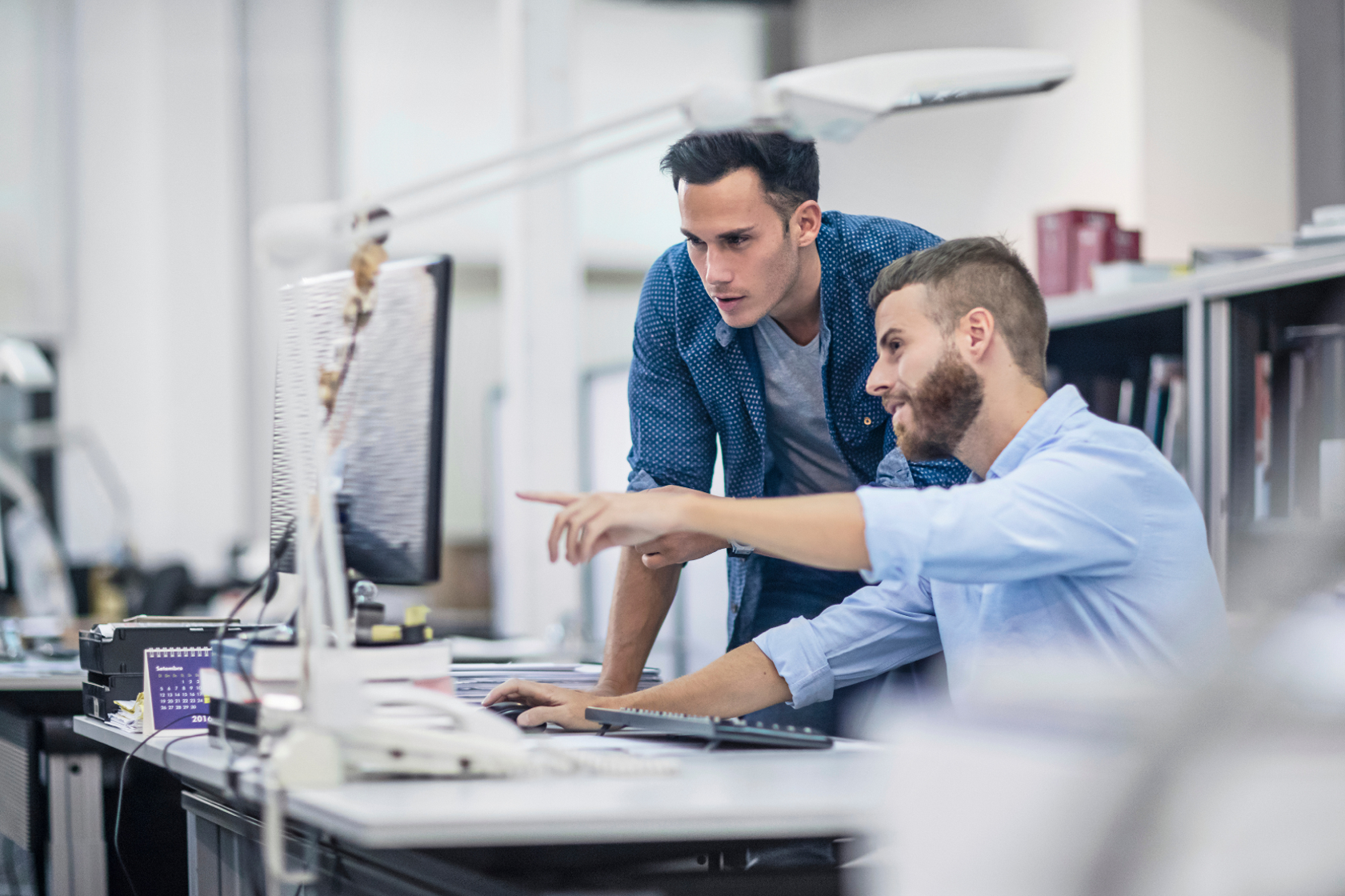 Zwei Männer in einem modernen Büro schauen konzentriert auf einen Computerbildschirm und diskutieren über Lichtplanung. Der eine sitzt und zeigt auf den Monitor, der andere steht daneben. Beide wirken konzentriert und in ihre Arbeit vertieft.