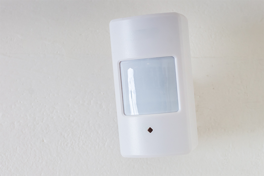Ein weißer Bewegungssensor, der an einer hellen Wand montiert ist und über ein rechteckiges, durchscheinendes Panel und eine kleine LED-Anzeige darunter verfügt.