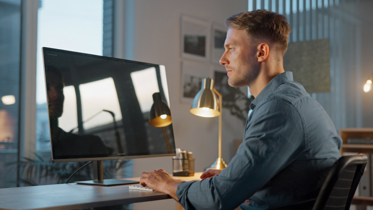 Ein Mann sitzt an einem Schreibtisch und arbeitet an einem Computer in einem modernen, gut beleuchteten Heimbüro mit einer Lampe, gerahmten Bildern an der Wand und einem großen Fenster, das den Blick auf den Bildschirmarbeitsplatz vermeidet.