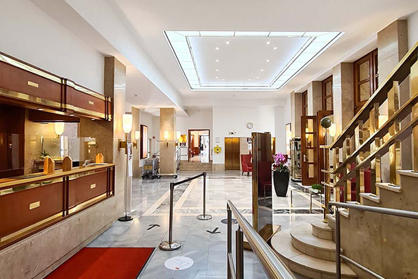 Eine gut beleuchtete Hotellobby mit modernen LED-Lichtsystemen, mit einem Check-in-Schalter auf der linken Seite, abgesperrten Warteschlangen für Kunden und einem roten Teppich, der zu den Schaltern führt. Auf der rechten Seite befindet sich eine große Treppe mit geschwungenem Design und Pflanzen entlang des gefliesten Bodens.