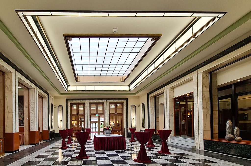 Ein geräumiger, eleganter Saal mit schwarz-weiß kariertem Marmorboden. Mehrere Stehtische mit roten Tischdecken sind über den Raum verteilt. Große Fenster und ein gitterförmiges Oberlicht, verstärkt durch strategische LED-Lichtsysteme, beleuchten den Raum, der mit dekorativen Wandelementen und Säulen ausgestattet ist.