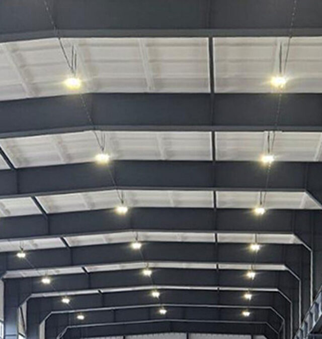 Ein Blick auf die Decke in einem großen Industrielager mit strukturierten Stahlträgern und gleichmäßig verteilten hellen Deckenleuchten.
