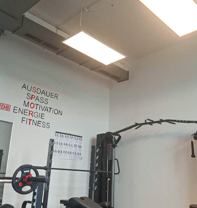 Ein Fitnessstudio mit Kraftgeräten, einer Bank und einer Wandbeschriftung mit der Aufschrift "AUSDAUER SPASS MOTIVATION ENERGIE FITNESS" auf Deutsch. Helle Deckenleuchten erhellen den modernen, sauberen Raum.