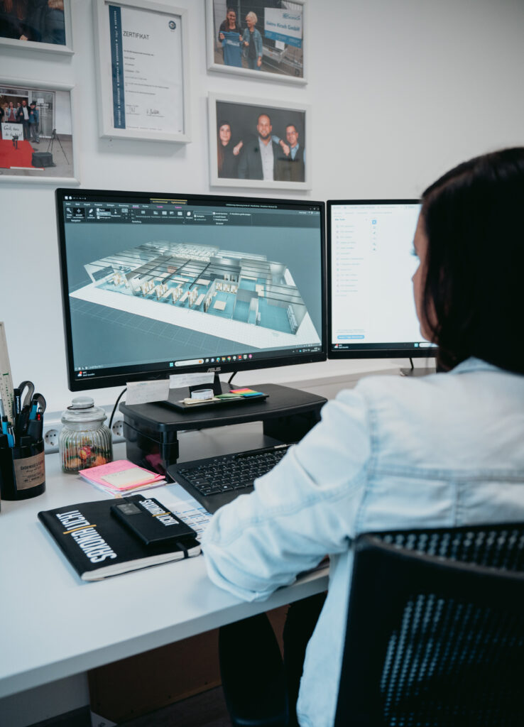 Eine Person sitzt an einem Schreibtisch mit zwei Monitoren und arbeitet an einem 3D-Architekturentwurf. Büromaterial, Bücher sowie gerahmte Zertifikate und Fotos schmücken den Arbeitsplatz.