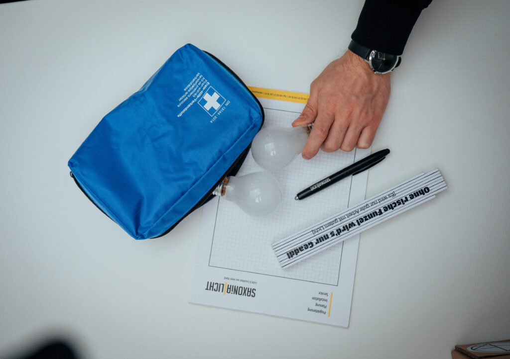 Die Hand einer Person ruht auf einem Tisch mit einer blauen Reißverschlusstasche, zwei Glühbirnen, einem schwarzen Stift, einem Lineal und einem Blatt mit der Überschrift „SENSOR LIGHT“ auf Millimeterpapier.