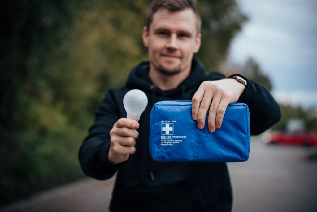 Ein Mann in einem schwarzen Kapuzenpullover hält in der einen Hand eine weiße Glühbirne und in der anderen einen blauen Erste-Hilfe-Kasten. Er steht im Freien vor einem unscharfen Hintergrund aus Bäumen und einer Straße.