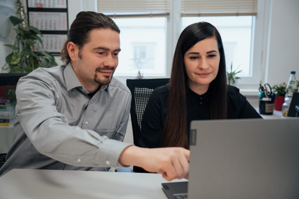 Ein Mann und eine Frau sitzen zusammen an einem Schreibtisch und schauen auf einen Laptop-Bildschirm. Der Mann zeigt auf den Bildschirm, und beide scheinen konzentriert zu sein. Im Hintergrund sind Büromaterial und Pflanzen zu sehen.