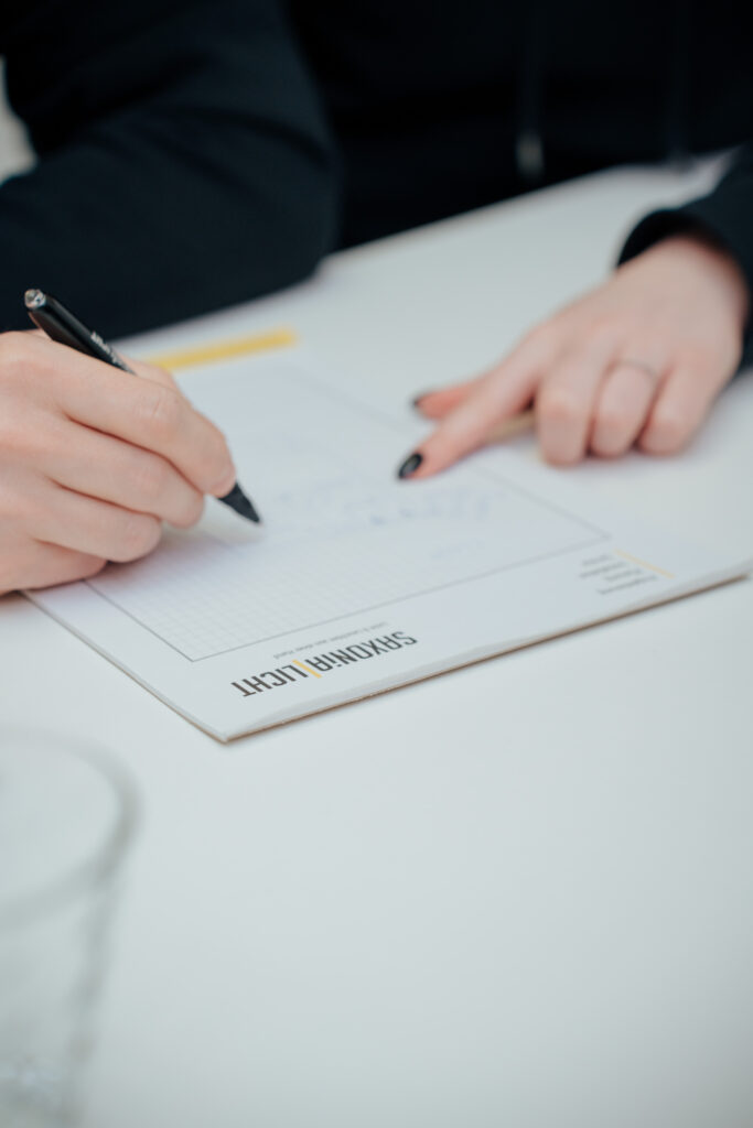 Eine Person schreibt auf einem Notizblock mit der Aufschrift „SIMONIN LIGHT“, während eine andere Hand mit schwarzem Nagellack auf das Papier zeigt. Beide Hände ruhen auf einem weißen Tisch.