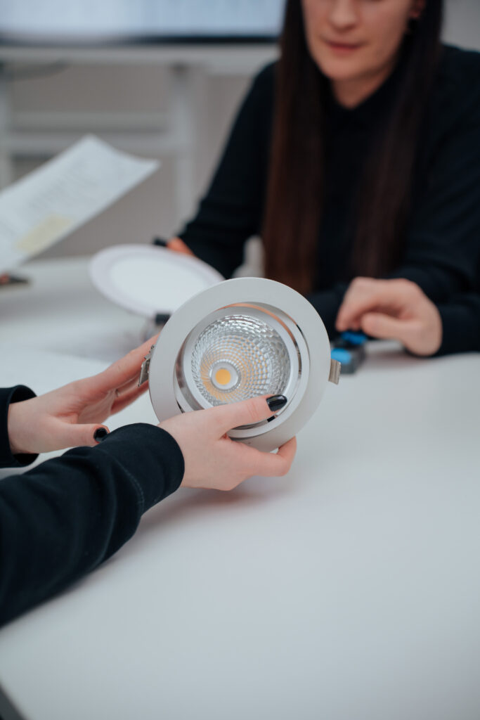 Zwei Personen sitzen an einem Tisch; eine hält eine kreisförmige LED-Einbauleuchte, während die andere, teilweise sichtbar, zusieht. Ein Blatt Papier und ein Stift liegen ebenfalls auf dem Tisch.
