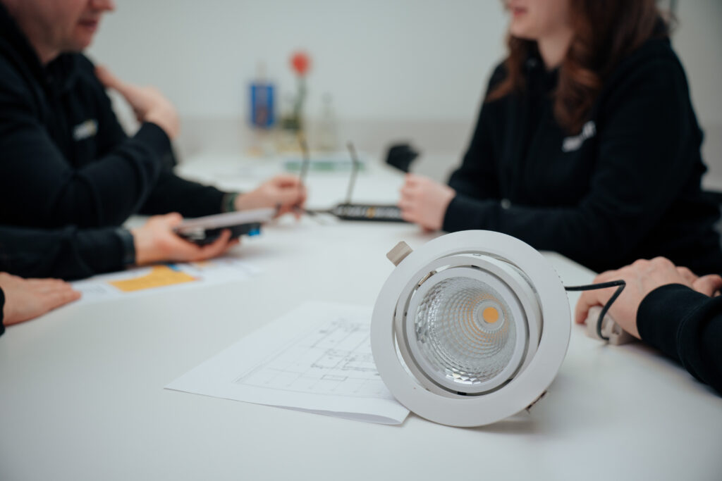 Eine Nahaufnahme einer runden LED-Deckenleuchte, die auf einem Papier mit Diagrammen ruht, während im Hintergrund drei Personen um einen weißen Tisch sitzen, sich unterhalten und Dokumente und Geräte in der Hand halten.