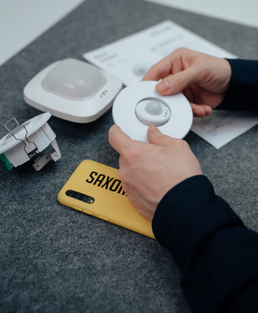 Eine Person hält ein rundes Gerät mit Bewegungssensor über eine graue Oberfläche, in deren Nähe sich ein gelbes Smartphone, Papiere und andere elektronische Komponenten befinden.