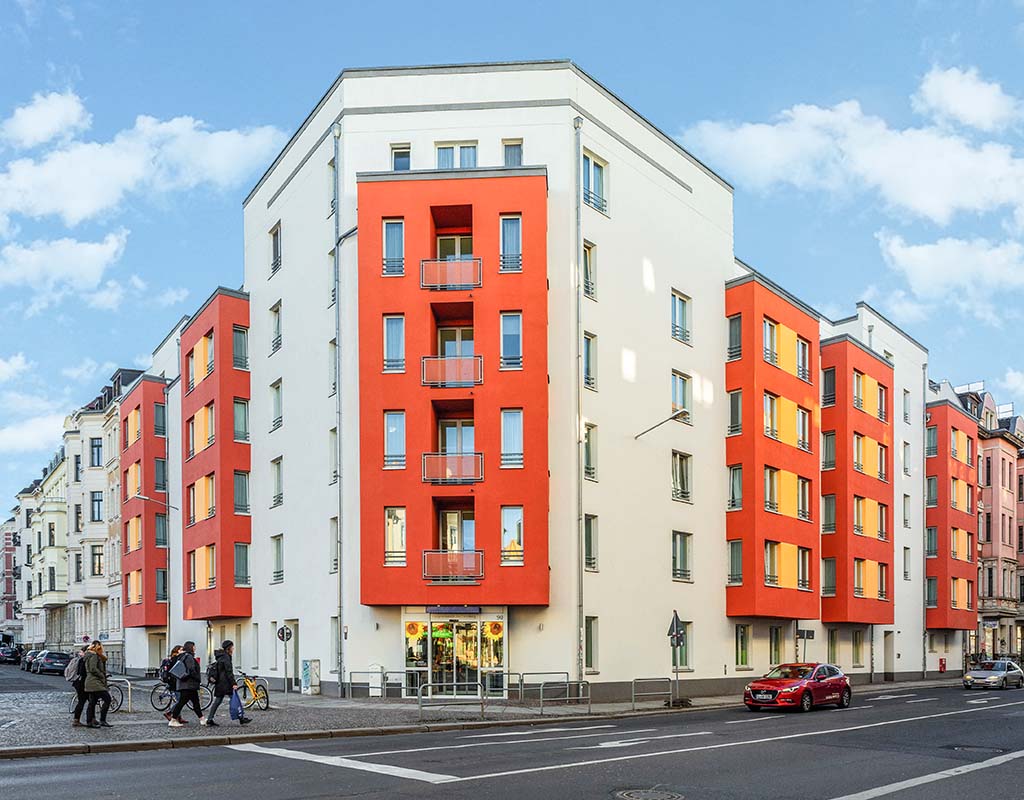 An einer Straßenecke steht ein modernes Wohnhaus mit rot-weißer Fassade. Das Gebäude wird von modernen LED-Beleuchtungssystemen beleuchtet und verfügt über mehrere Balkone und große Fenster. An der Seite parkt ein rotes Auto, und in der Nähe sind Menschen zu Fuß oder mit dem Fahrrad unterwegs. Im Hintergrund sind umliegende Gebäude zu sehen.
