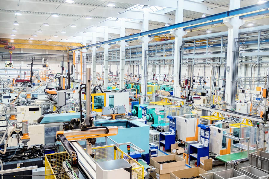 Im Inneren einer großen, gut beleuchteten Fabrik werden moderne Beleuchtungsindustriehallen mit zahlreichen Maschinen und Geräten präsentiert. Der Raum ist organisiert und umfasst Fließbänder, Verkabelungen und Lagerbereiche. Hohe Decken und Fenster lassen natürliches Licht in die geräumige Industrieumgebung strömen.