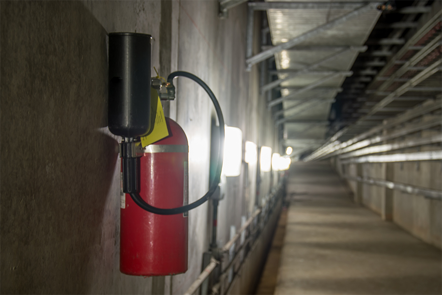 Ein roter Feuerlöscher ist an einer Betonwand in einer langen, industriellen Halle mit hellen Lichtern an der Wand und Metallgittern über dem Kopf angebracht.