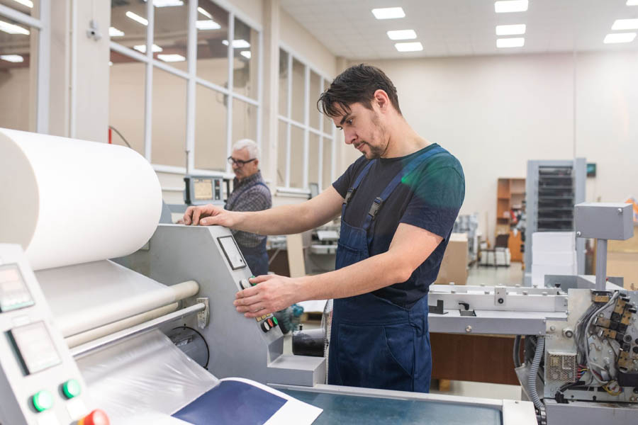 Ein Mann im blauen Overall bedient eine Druckmaschine und justiert Bedienelemente auf einem Bedienfeld. Im Hintergrund ist ein weiterer Mann zu sehen. Sie befinden sich in einer gut beleuchteten Industrieumgebung und profitieren von der effizienten „Beleuchtung Industriehallen“ mit großen Fenstern und umgebender Ausrüstung.