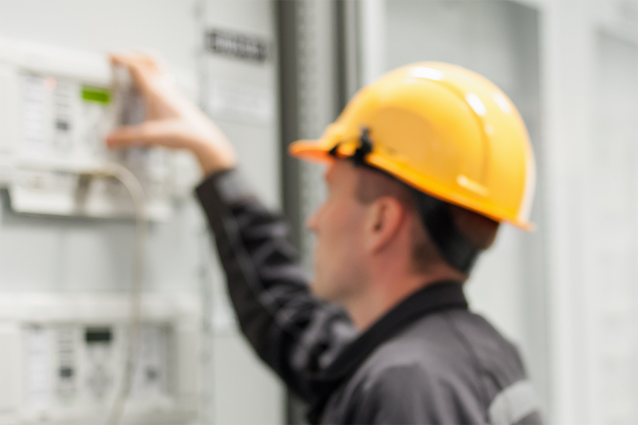Eine Person, die einen gelben Schutzhelm und dunkle Arbeitskleidung trägt, prüft oder bedient elektrische Schalttafeln; das Bild ist unscharf, so dass Details unklar sind.