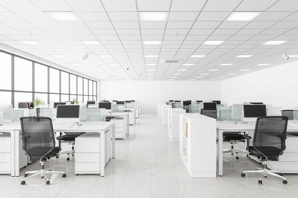 Ein geräumiges Großraumbüro mit Reihen weißer Schreibtische und ergonomischen schwarzen Stühlen. Jeder Schreibtisch ist mit einem Computermonitor und Büromaterial ausgestattet. Große Fenster auf der linken Seite sorgen für natürliches Licht, während die Decke mit LED-Technik-Beleuchtung für optimale Helligkeit ausgestattet ist.
