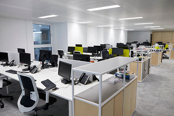 Ein modernes Großraumbüro mit Reihen von leeren Schreibtischen, die jeweils mit Computern, Telefonen und Bürostühlen ausgestattet sind. Der Raum ist mit Oberlichtern gut beleuchtet und hat weiße Wände und große Fenster.