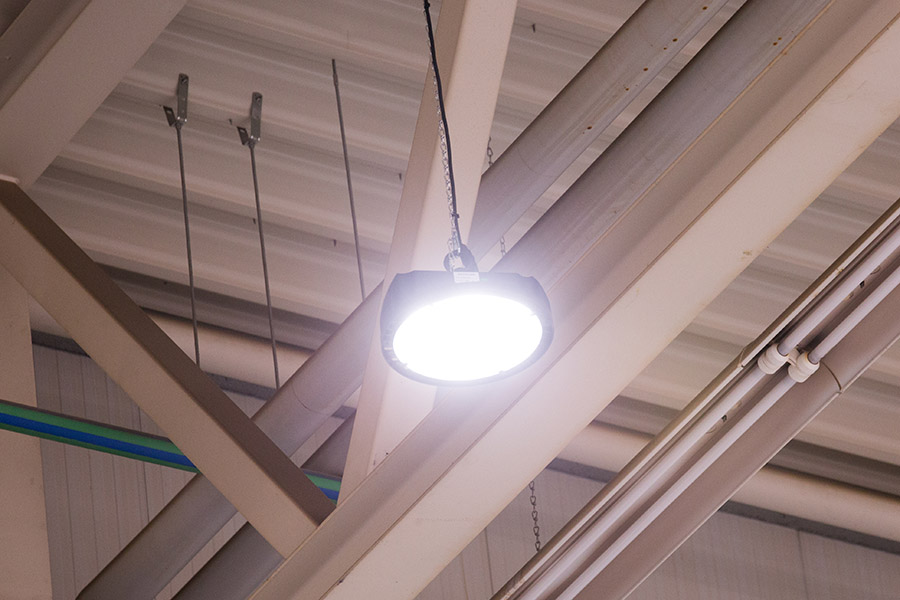 Eine helle industrielle Deckenleuchte erhellt die Umgebung und wirft einen Schein auf die darüber liegenden Metallträger und Rohre. Dieses Design von Beleuchtung Industriehallen hebt die strukturellen Elemente hervor und betont sowohl Form als auch Funktion in Räumen mit hohen Decken.
