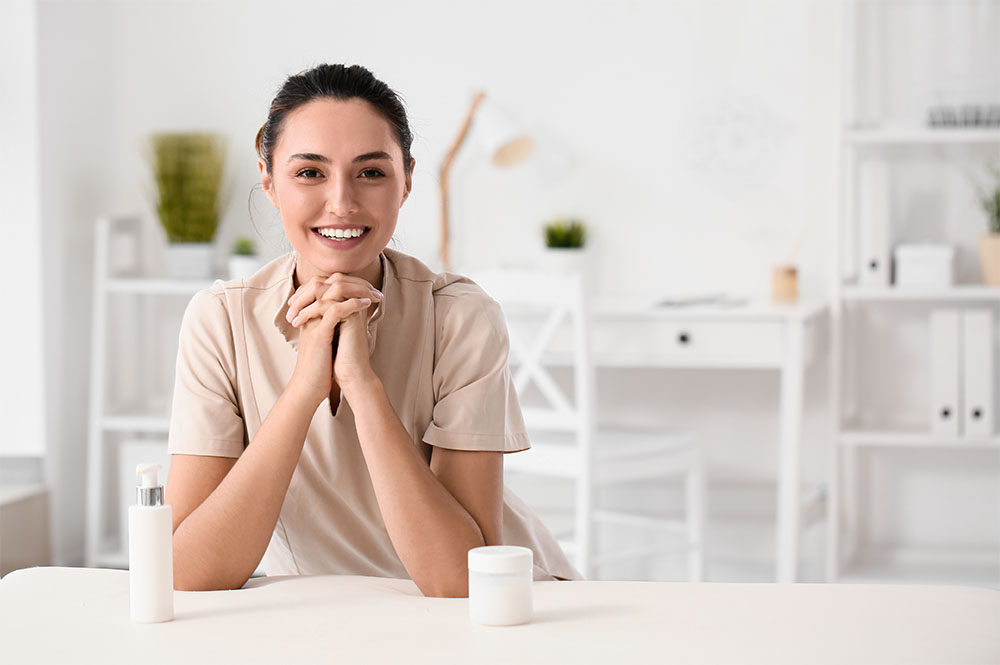 Eine lächelnde Frau sitzt mit verschränkten Händen an einem Tisch, neben sich eine weiße Flasche und ein weißes Glas. Der Hintergrund zeigt einen hellen, modernen Raum mit Regalen, Pflanzen und einem Schreibtisch.
