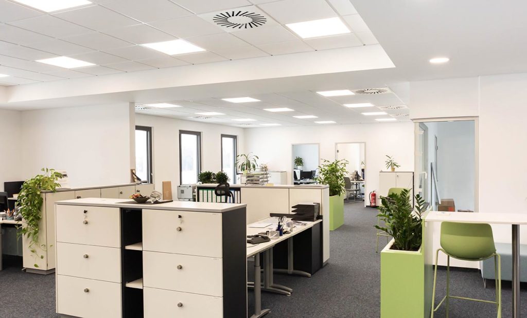 Geräumiges, modernes Büro mit weißen Wänden, Deckenleuchten und großen Fenstern. Ausgestattet mit Schreibtischen, Aktenschränken und grünen Akzenten durch Pflanzen und Stühle. Der Raum ist organisiert und gut beleuchtet mit modernen LED-Beleuchtungssystemen, verschiedenen Arbeitsplätzen und einer sauberen, professionellen Atmosphäre.