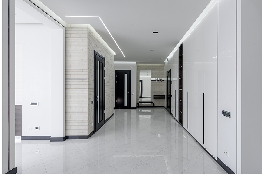 Moderner, minimalistischer Flur mit glänzend weißen Bodenfliesen, schlichten weißen Wänden, schwarzen Akzenten, eingebautem Stauraum und eingebauter Deckenbeleuchtung. Im Hintergrund sind mehrere Türen und eine kleine Bank zu sehen.