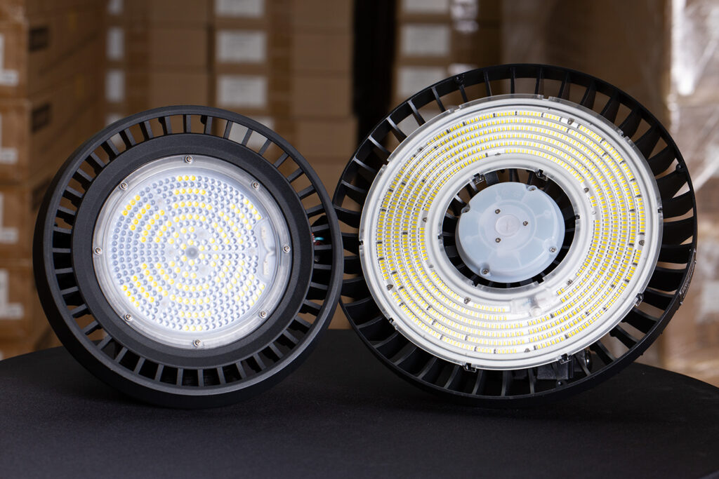 Zwei runde industrielle LED-Hallenstrahler auf einer schwarzen Oberfläche, wobei ein Strahler nach vorne zeigt und der andere sein LED-Array zeigt. Kartons und Regale sind im Hintergrund unscharf.