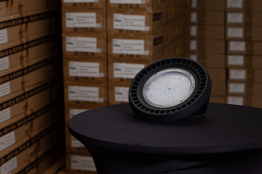 Auf einem runden, schwarz gedeckten Tisch steht eine runde LED-Industrieleuchte in einem schwarzen Gehäuse, im Hintergrund stapeln sich Kartons.