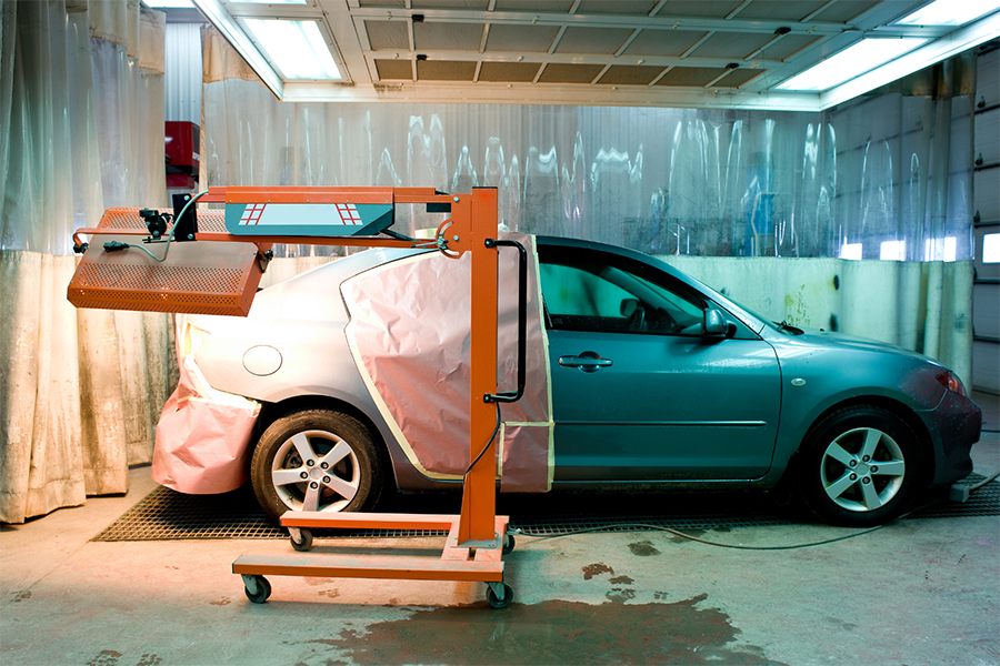 Ein blaues Auto mit abgeklebten Scheiben und abgeklebten Bereichen steht in einer Autowerkstatt. Neben dem Auto steht eine orangefarbene Infrarot-Härtungslampe, vermutlich zum Trocknen von Lack. Die Werkstatt ist mit Plastikvorhängen und Leuchtstoffröhren ausgestattet.