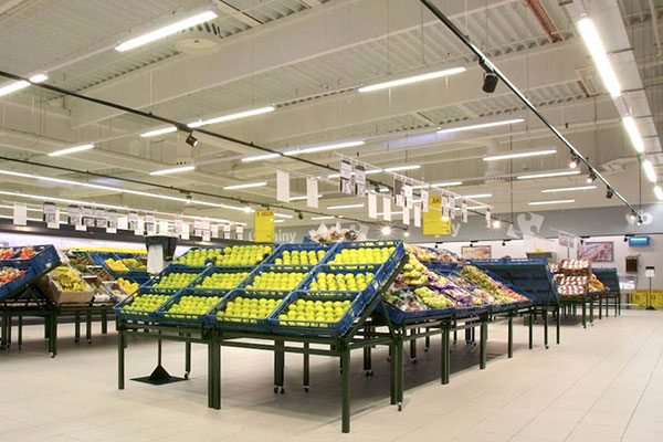 Eine geräumige Obst- und Gemüseabteilung im Supermarkt mit ordentlich gestapeltem frischem Obst und Gemüse auf mehreren Verkaufsregalen, die von LED-Beleuchtungssystemen beleuchtet werden. Äpfel, Birnen und andere Produkte sind attraktiv angeordnet und bieten eine saubere und organisierte Einkaufsumgebung.