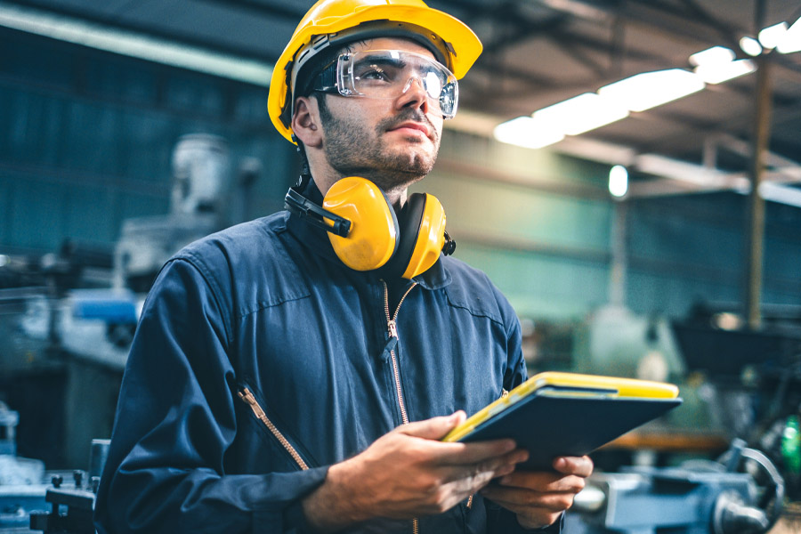 Ein Arbeiter in blauem Overall und gelbem Bauhelm steht in einer Fabrik, die von der präzisen Beleuchtung Industriehallen beleuchtet wird. Er trägt Schutzbrille und Ohrenschützer und hält ein Tablet in der Hand. Im unscharfen Hintergrund sind Maschinen zu sehen, die das geschäftige Industrieumfeld hervorheben.