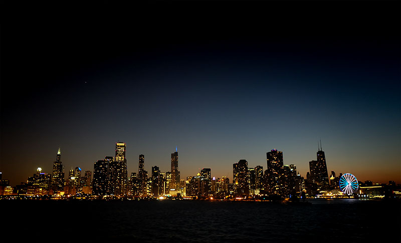 Eine Skyline bei Nacht mit hell erleuchteten Gebäuden vor einem dunkelblauen Himmel. Das Bild zeigt auf der rechten Seite ein beleuchtetes Riesenrad mit LED-Beleuchtungssystemen, und im Vordergrund sieht man die Spiegelung der Lichter der Stadt auf dem Wasser.