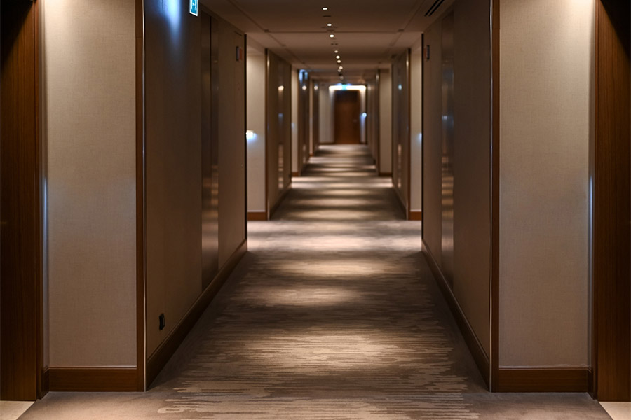 Ein moderner, gut beleuchteter Hotelflur mit beigen Wänden, Teppichboden und Holztürrahmen. Sicherheitsbeleuchtung im Unternehmen sorgt dafür, dass Deckenleuchten ein Muster aus Licht und Schatten entlang des Flurs erzeugen, der zu einer Tür am Ende führt.