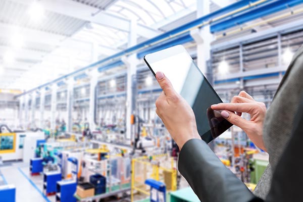 Eine Person hält ein Tablet und bedient es in einer hell erleuchteten, weitläufigen Produktionshalle voller Maschinen und Geräte. Die Hand der Person ist im Fokus, während die industrielle Umgebung, die durch moderne LED-Beleuchtungssysteme aufgewertet wird, leicht verschwommen erscheint.