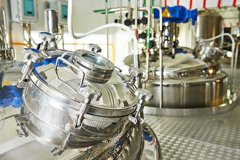 Glänzende Edelstahltanks mit runden Deckeln stehen auf einem Metallgitterboden in einer großen sterilen Produktionsanlage, die wahrscheinlich für chemische, pharmazeutische oder Lebensmittelverarbeitungszwecke verwendet wird. Verschiedene Rohre und Messgeräte sind angeschlossen, alle von modernen LED-Beleuchtungssystemen beleuchtet.