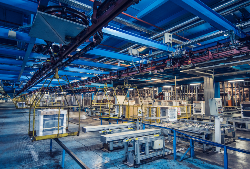 Ein modernes Fabrikinterieur mit Förderbändern, Metallgerüsten und aufgehängten Maschinen, die große Geräte verarbeiten - hervorgehoben durch eine präzise Lichtplanung für eine optimale Beleuchtung in dieser industriellen Umgebung.